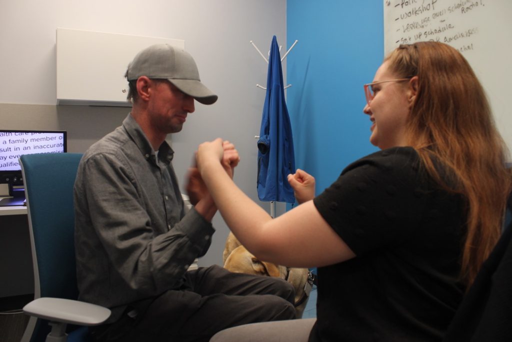 Ryan Ollis, Deafblind Coordinator & Rachel Kavanagh, DeafBlind Intervenor sit across from each other in an office and communicate using tactile ASL. Ryan wears a grey hat and grey shirt, while Rachel wears a black shirt; they are both smiling. In the background to the left, a CCTV shows magnified text, and the head of a guide dog is seen near Ryan’s knee.