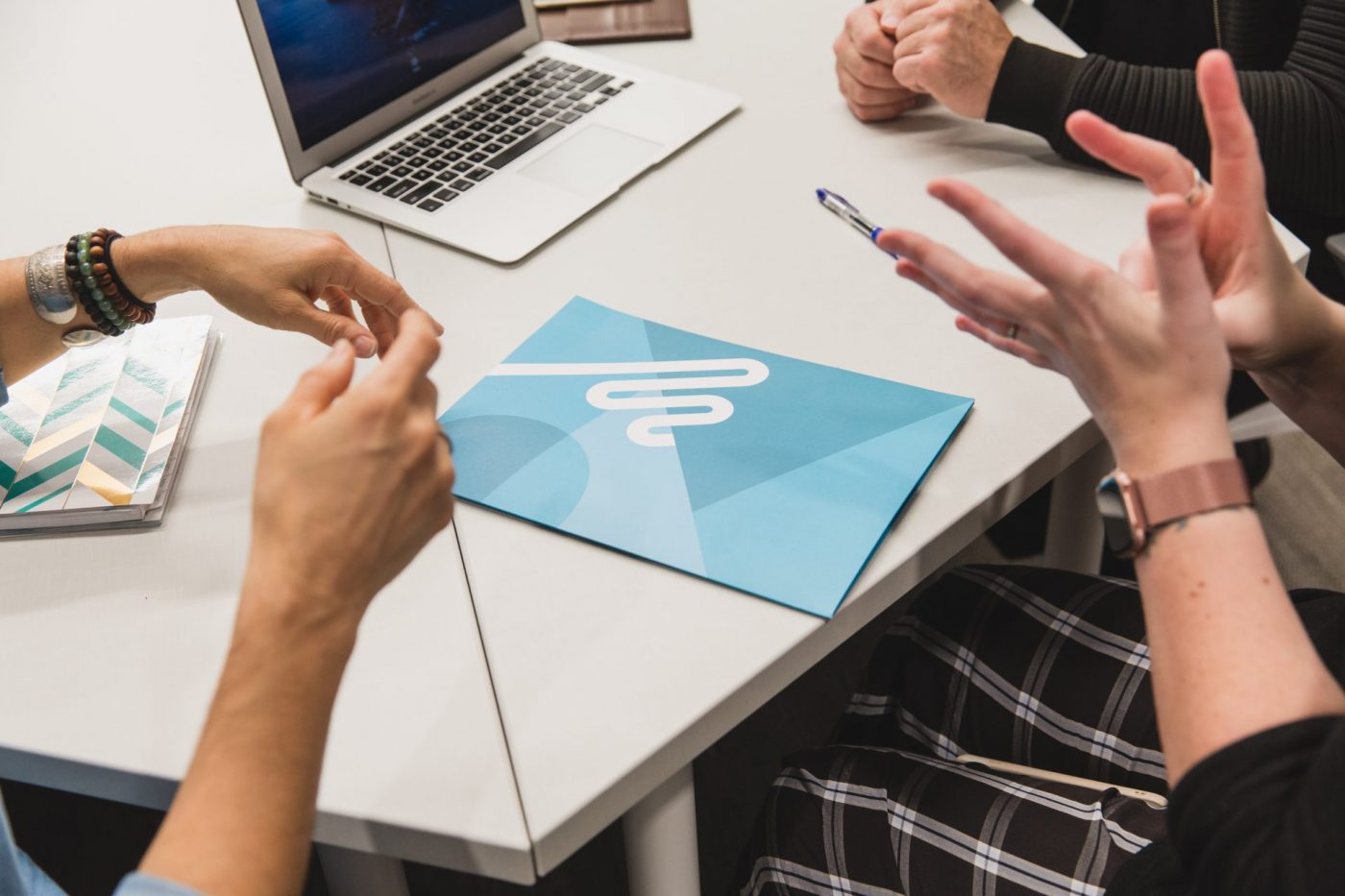 A closer shot of the hands of three people putting on a white desk and signing to each other. A blue brochure with Wavefront Centre logo is on the table in the middle of the photo. A laptop shows on the farer top of the photo.