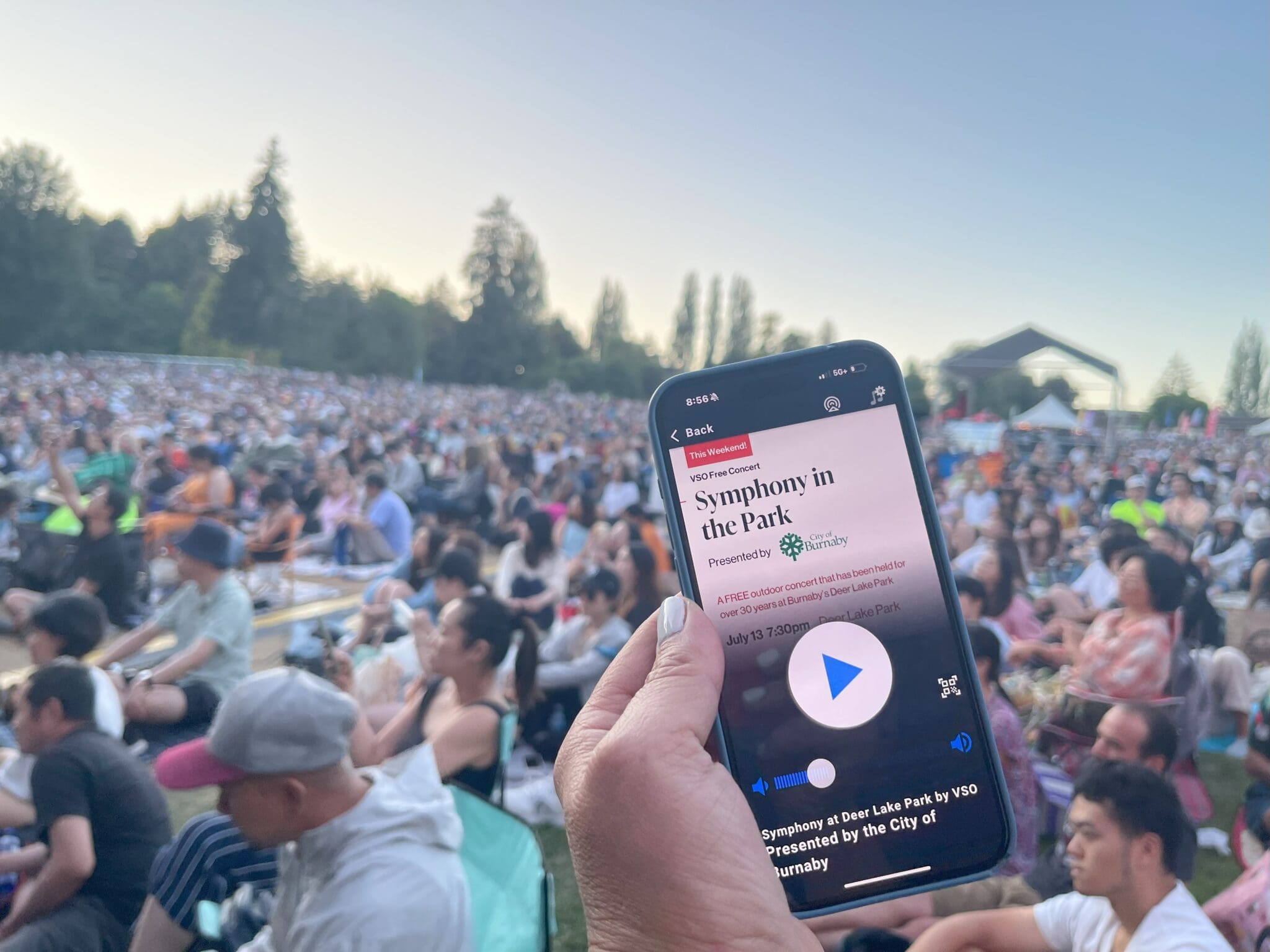 A person holds a smartphone displaying information about a "Symphony in the Park" event while attending an outdoor concert. The phone screen shows event details, including that it is a free concert presented by the City of Burnaby. In the background, a large crowd sits on blankets and chairs, attentively watching the performance at a park. The stage and trees are visible in the distance under a clear evening sky.