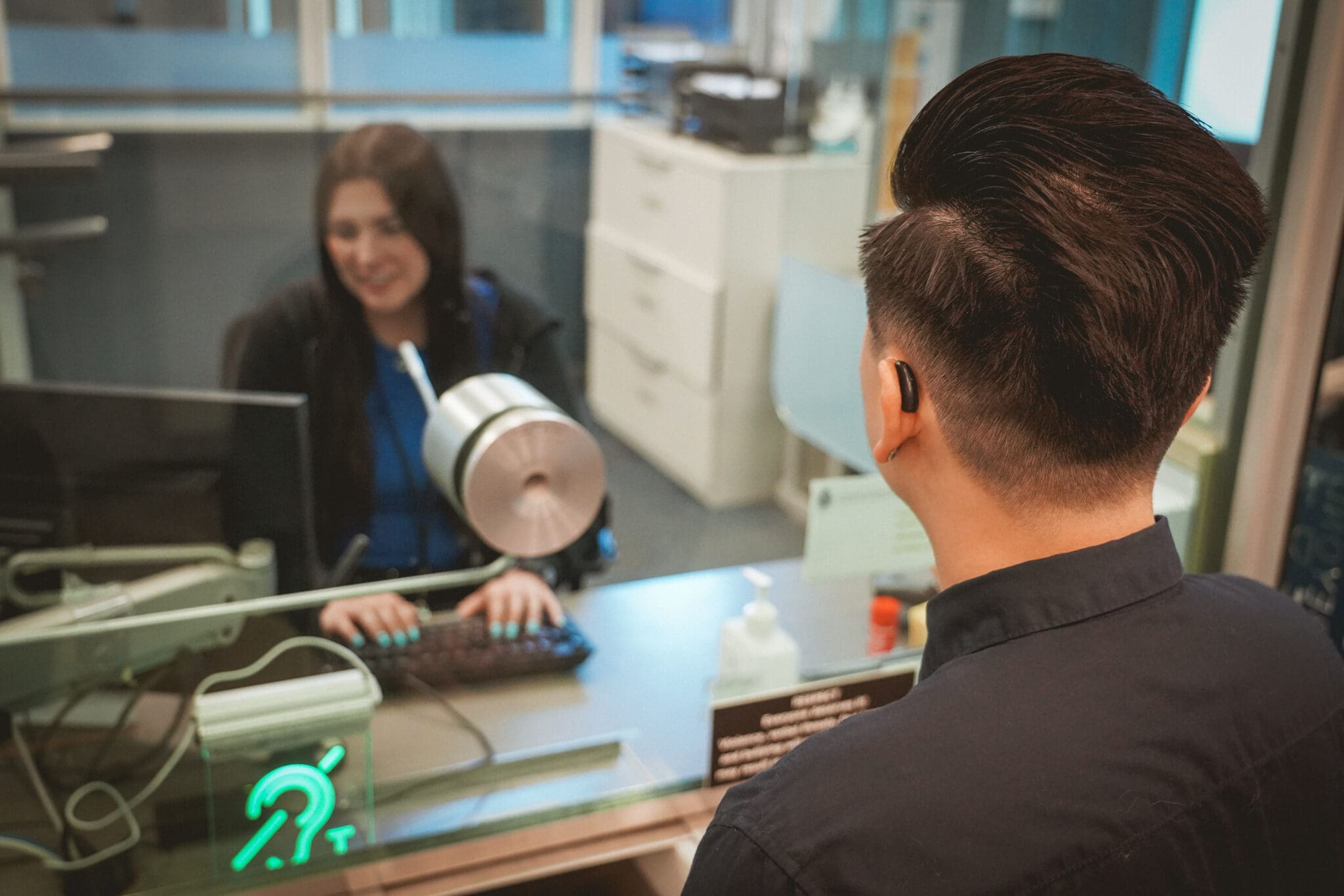 A man wearing a hearing aid communicates with a receptionist through a service counter equipped with a hearing loop system. The green illuminated hearing loop sign is visible on the glass barrier, indicating assistive listening technology is available. The receptionist smiles while typing on a keyboard, and a microphone device is positioned between them to support clear communication.
