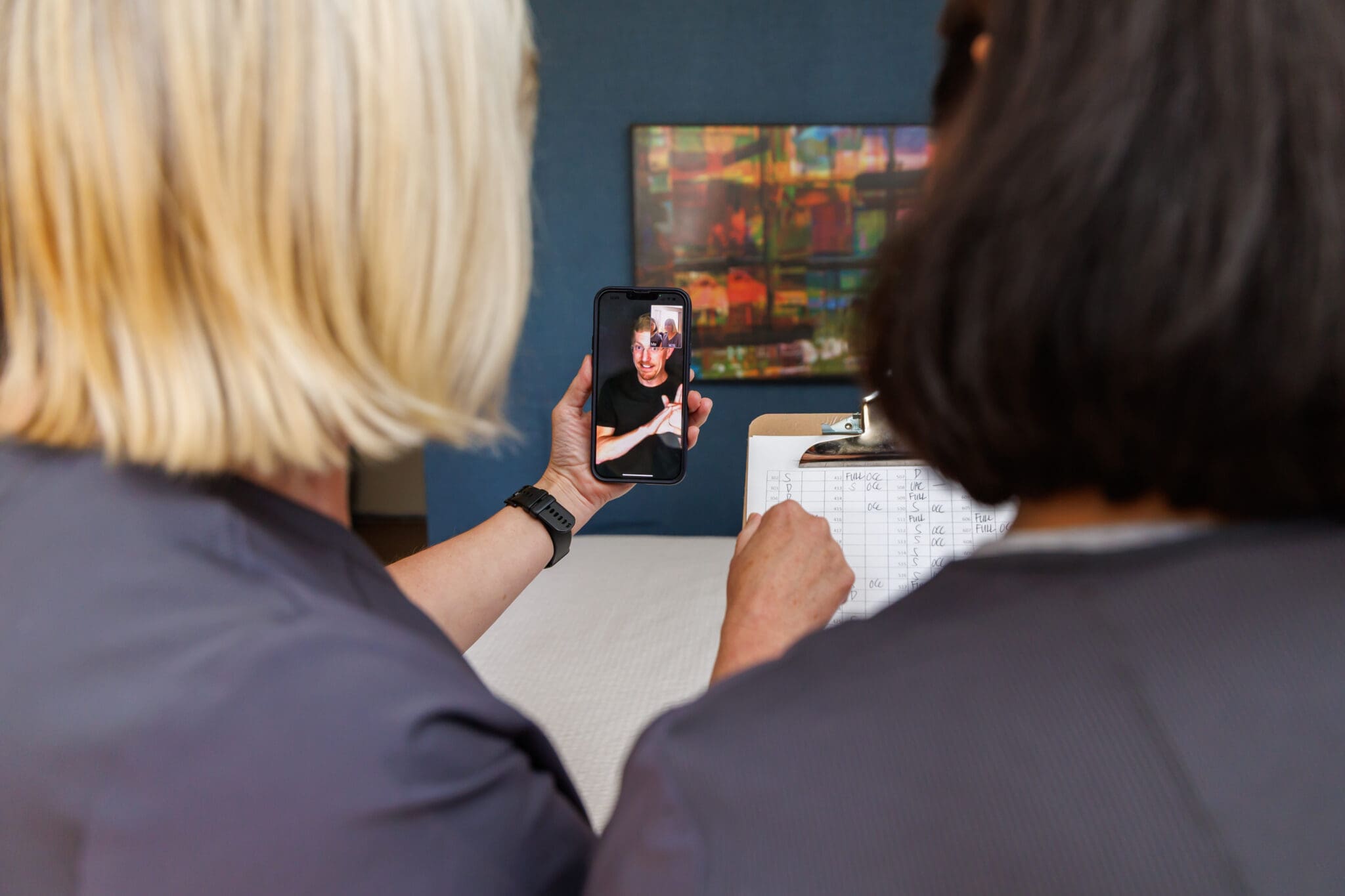 Two people are seen from behind, one holding a smartphone showing a live video of a sign language interpreter, the other holding a clipboard with notes. The image captures a moment from a Wavefront Centre and Holiday Inn Express Metrotown field test of Virtual On-Demand Interpreting, enabling real-time communication between Deaf and hearing individuals without advance booking of an interpreter.