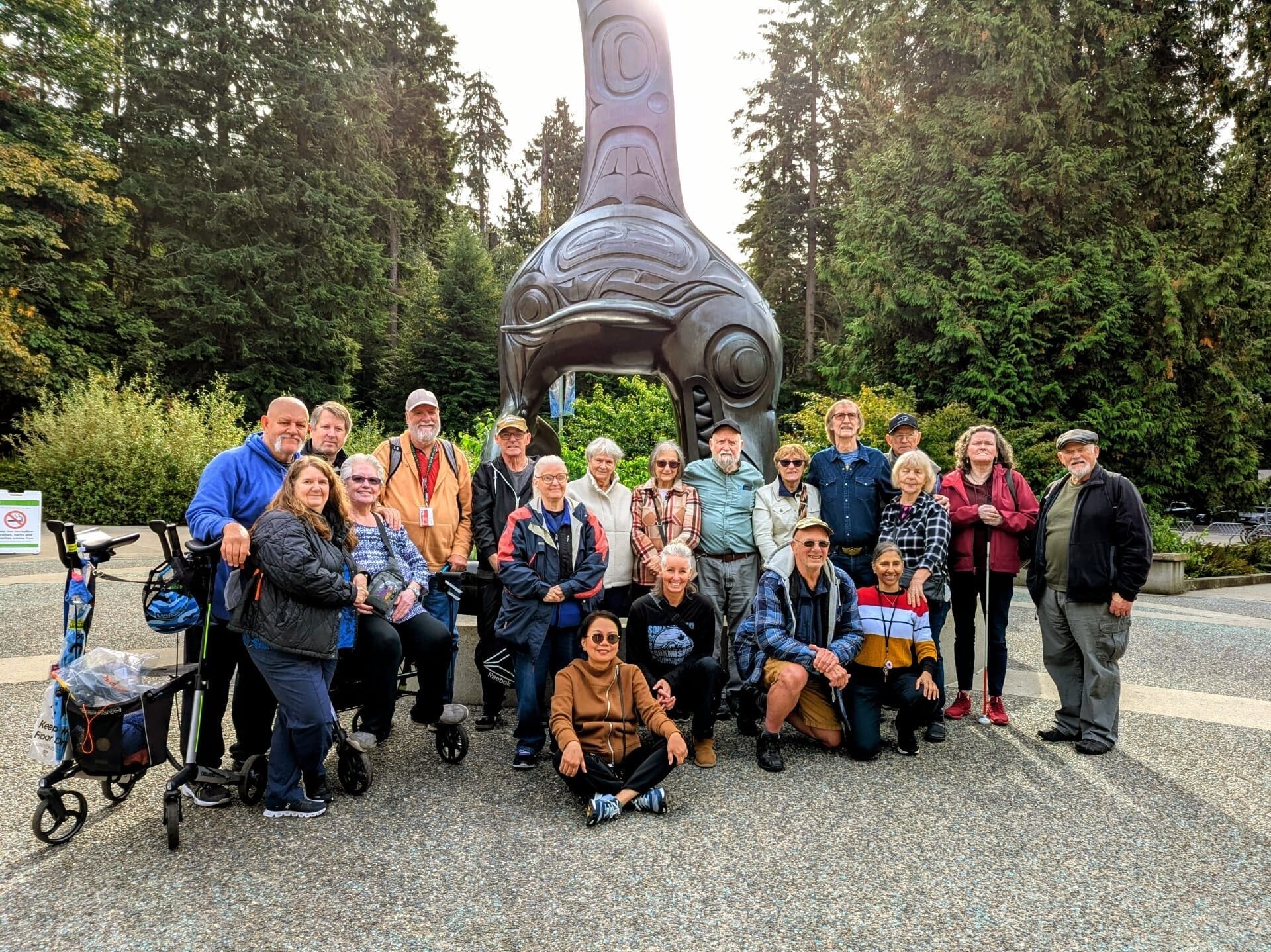 A group of Deaf, DeafBlind, and Hard of Hearing seniors pose together outdoors in front of a large orca sculpture at the Vancouver Aquarium. The group is smiling and dressed for cool weather, surrounded by trees and natural greenery. This photo was taken during Wavefront Centre’s Seniors’ Appreciation Day outing as part of the Better at Home program.