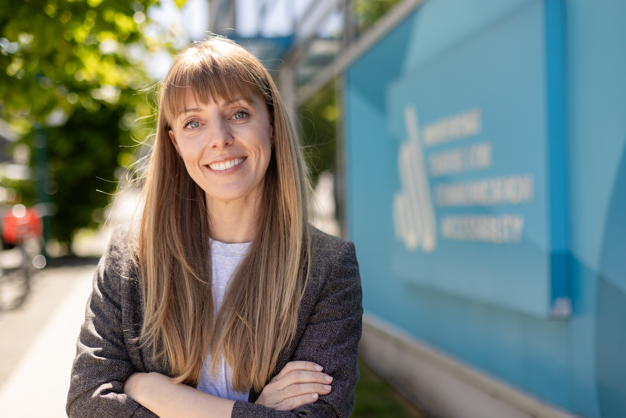 Smiling woman with long hair stands outdoors near a sign reading "Wavefront Centre for Communication Accessibility." Sunlight and greenery in the background.
