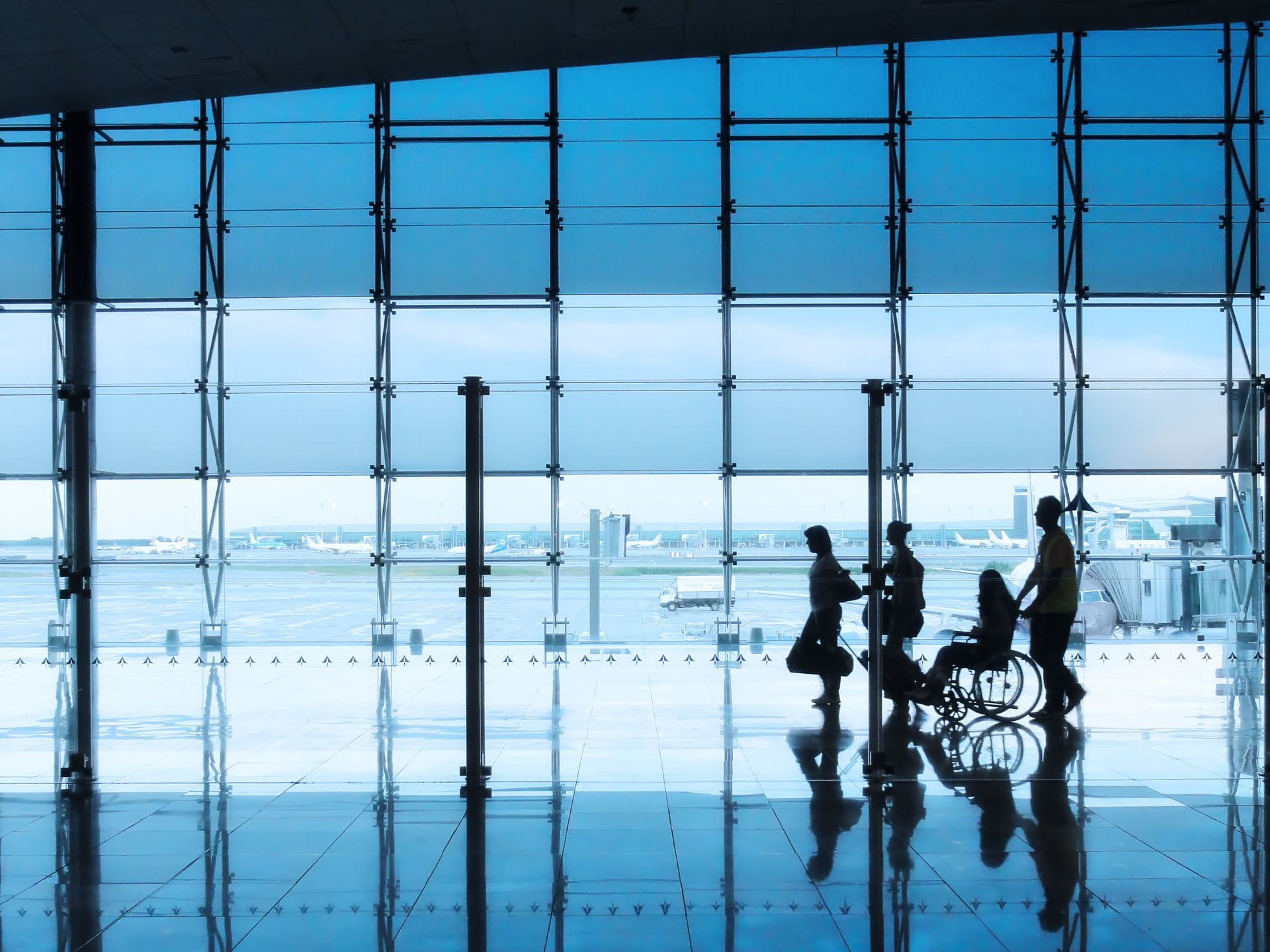 Silhouettes of travelers, including someone in a wheelchair, move through a bright airport terminal with large glass windows showing the runway.