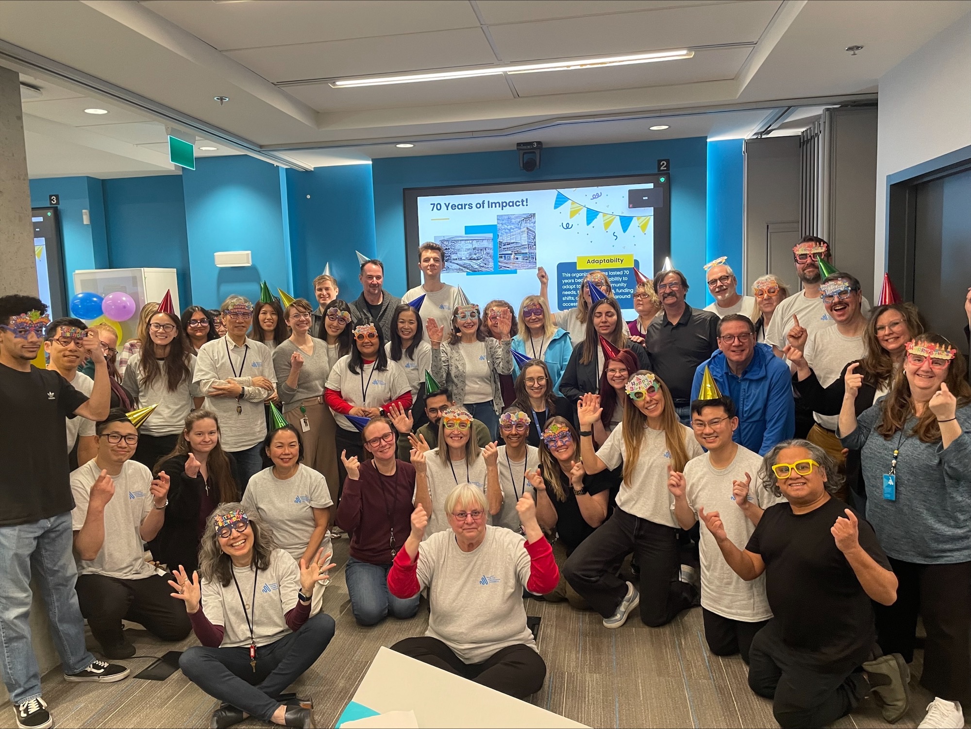 A joyful group of people, many wearing party hats and glasses, gather in a brightly lit room. A screen behind them shows "70 Years of Impact!" Balloons add a festive atmosphere.