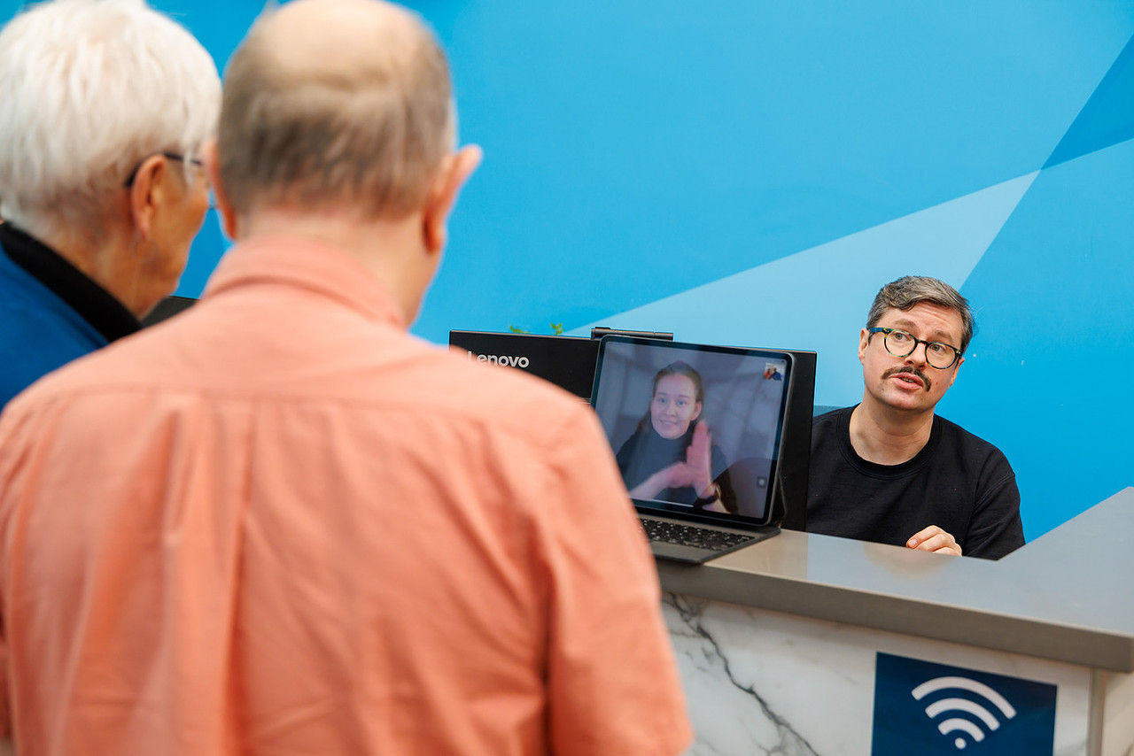 A large group of Deaf, DeafBlind, and Hard of Hearing seniors gathered in a bright conference room at Wavefront Centre, wearing pink in support of Pink Shirt Day. They are smiling and making heart shapes with their hands, symbolizing kindness and inclusion. The room has blue walls, modern lighting, and tables arranged for a social event.