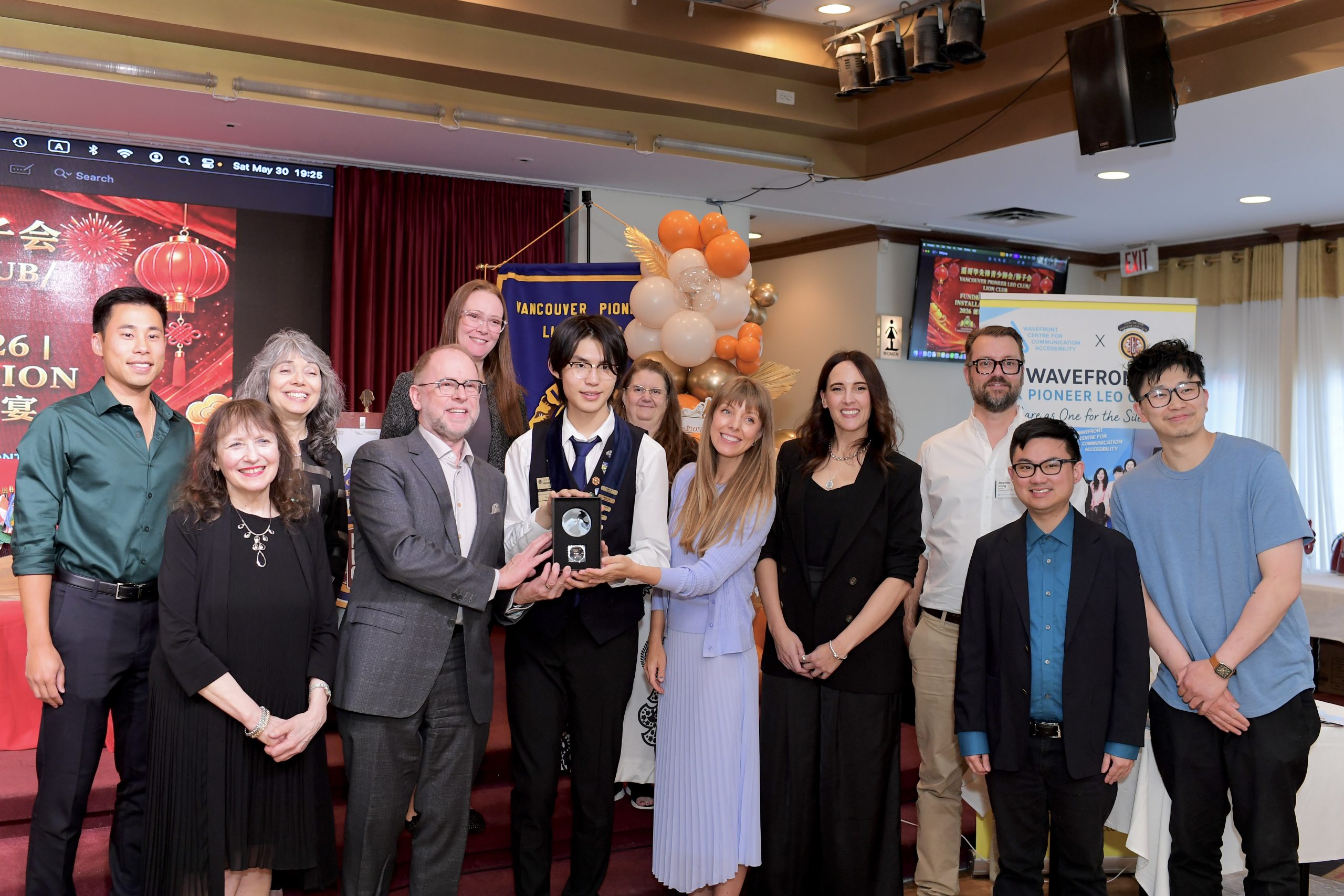  A group of Wavefront Centre staff stands smiling with Vancouver Pioneer LEO Club President Alex Zhang, after being presented with an award. Balloons and banners are in the background, conveying a celebratory event.
