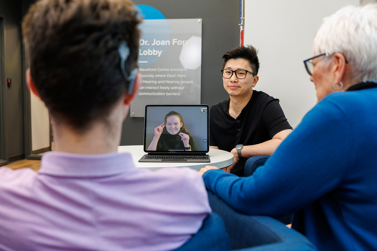 A person uses a tablet at a reception desk to connect with an ASL-English interpreter through Access ASL, while a staff member assists nearby.