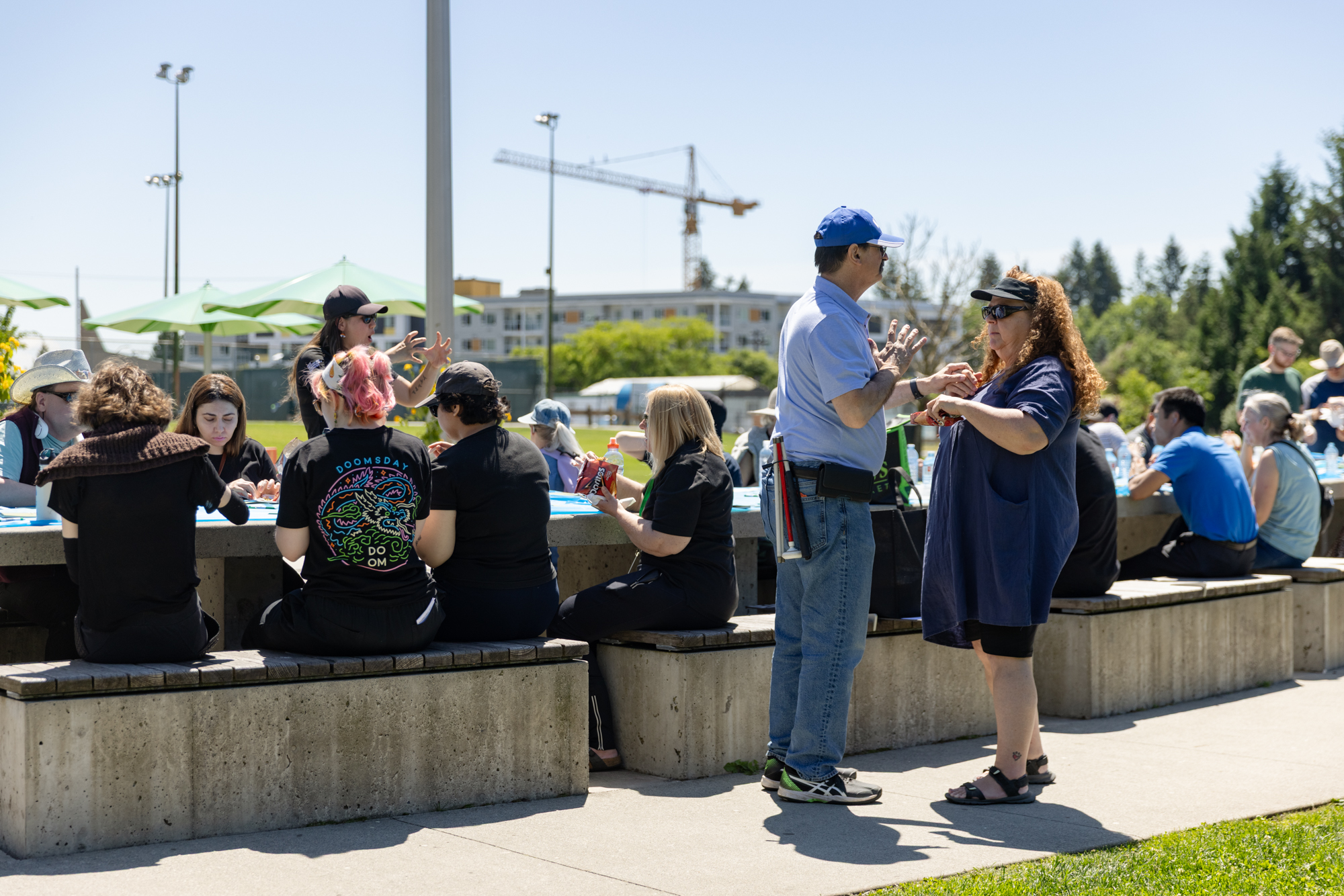 A man and a woman communicating through tactile sign language stand in the foreground during the DeafBlind Awareness Picnic at Lafarge Lake in Coquitlam, BC. Behind them, participants sit and socialize around outdoor picnic tables while enjoying lunch and activities on a sunny day. 