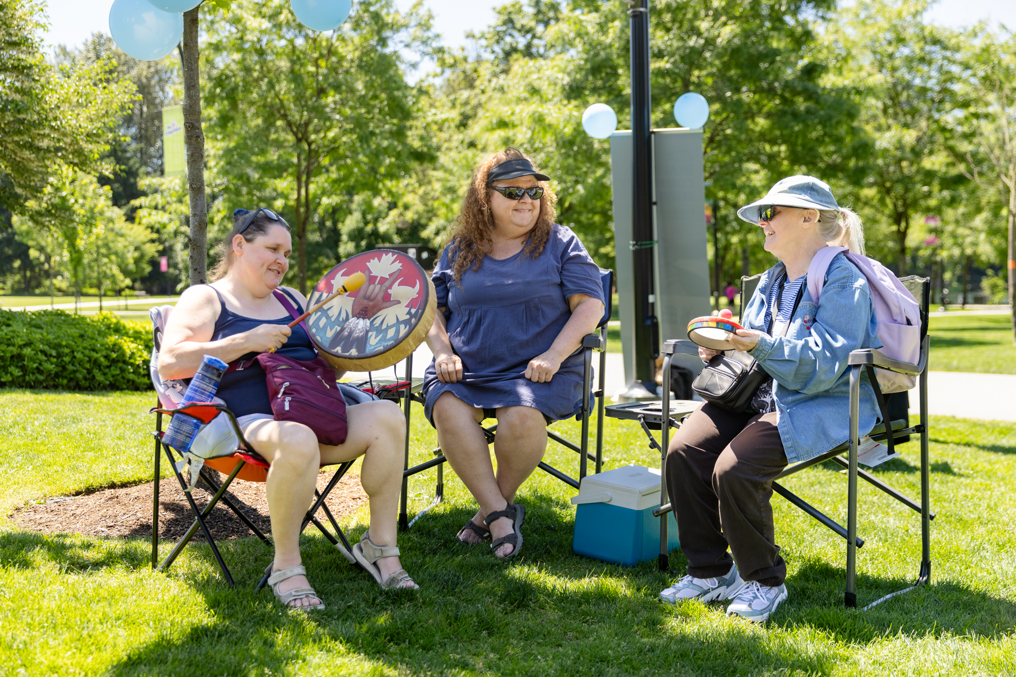 Three women sit together on lawn chairs in a shaded area during the DeafBlind Awareness Picnic at Lafarge Lake in Coquitlam, BC. One participant holds a hand drum and drumstick while another holds a small percussion instrument. The group smiles and engages in conversation on a sunny day surrounded by trees and green space.