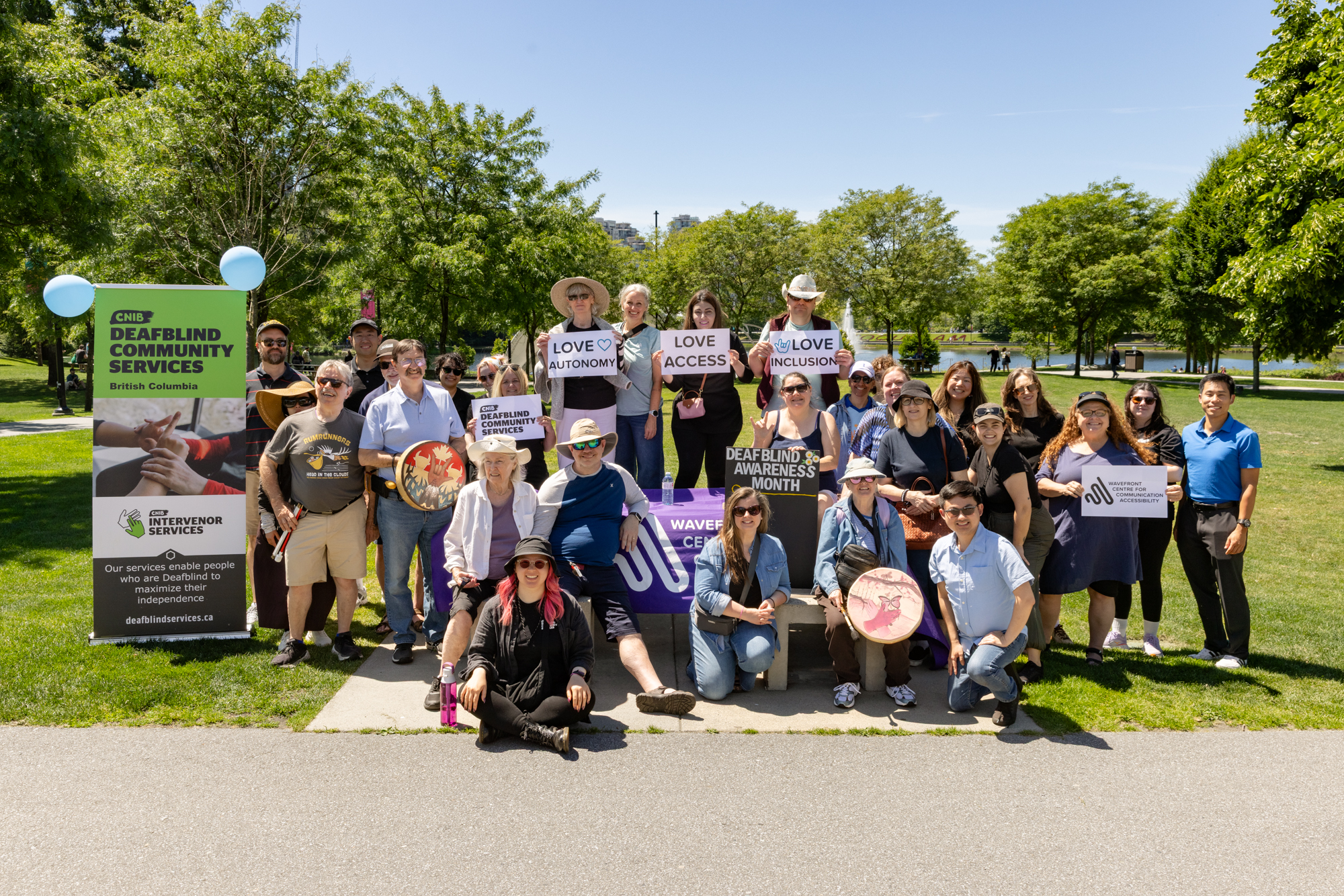 Group photo of approximately 30 participants, staff, volunteers, and community partners gathered outdoors at Lafarge Lake in Coquitlam, BC, during the DeafBlind Awareness Picnic on June 12, 2026. Attendees stand and sit around Wavefront Centre and CNIB Deafblind Community Services signage, holding signs that read "Love Access," "Love Inclusion," "Love Autonomy," and "DeafBlind Awareness Month." Trees, green space, and the lake are visible in the background on a sunny day.