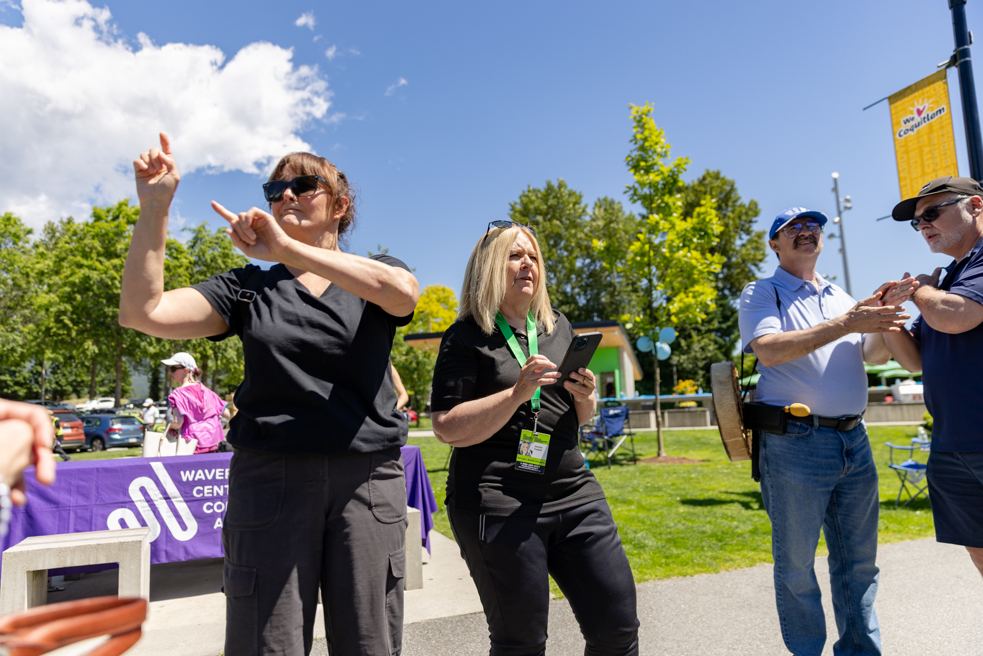 At the DeafBlind Awareness Picnic in Coquitlam, BC, a woman stands holding a phone while reading the Province of British Columbia's Royal Proclamation for DeafBlind Awareness Month. Beside her, an interpreter signs the message for attendees. In the foreground, two participants communicate using tactile sign language, with one person placing their hands over the other's to receive information through touch. A Wavefront Centre banner and picnic activities are visible in the background on a sunny day.