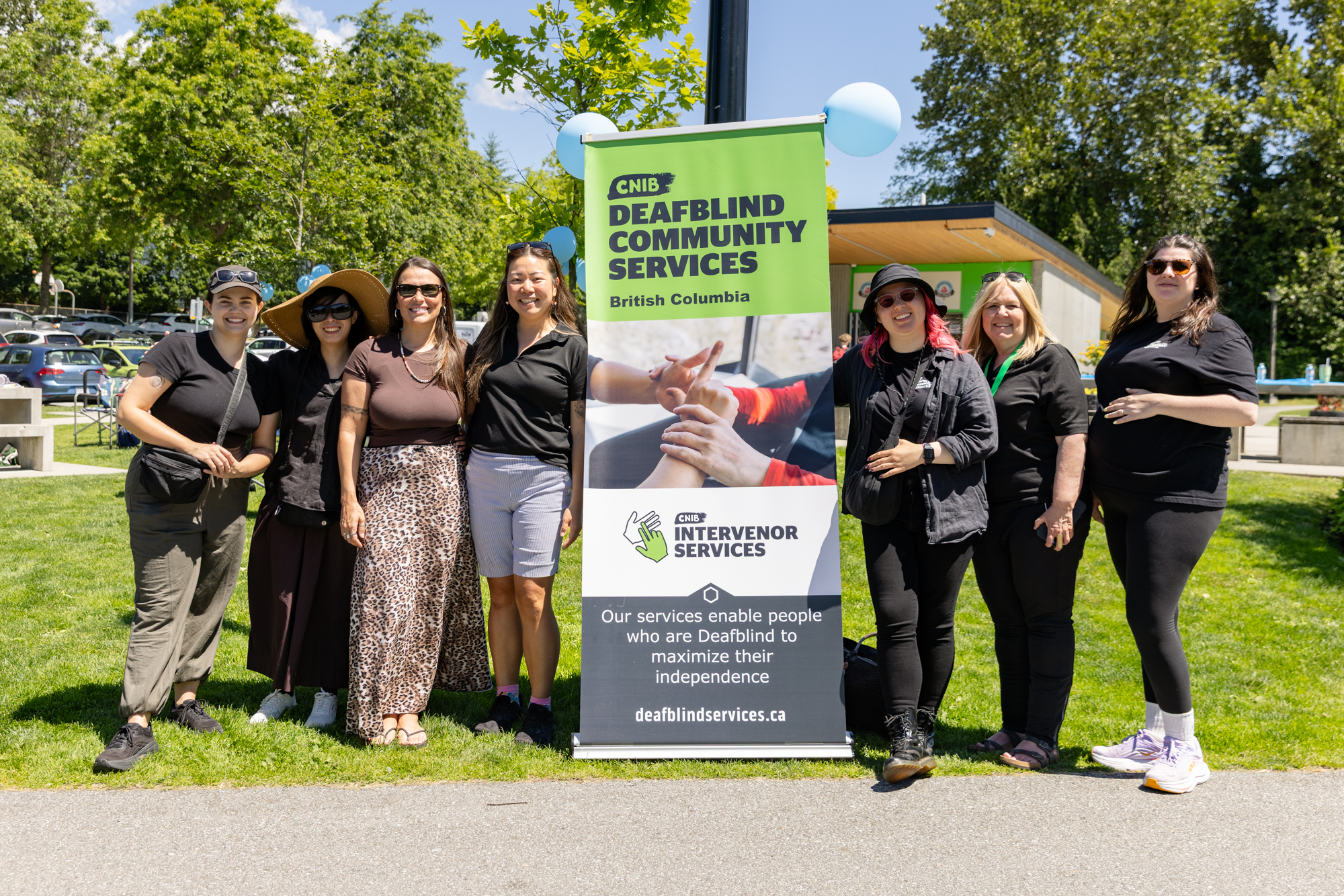 Seven staff members from CNIB Deafblind Community Services BC stand together outdoors beside a CNIB Deafblind Community Services banner at the DeafBlind Awareness Picnic in Coquitlam, BC. The group smiles for a photo on a sunny day surrounded by trees and green space. 