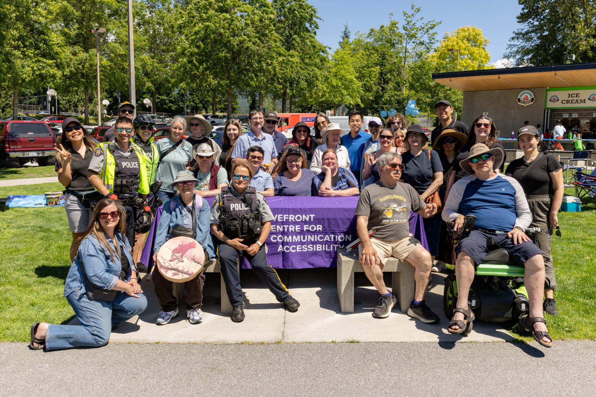 A large group of DeafBlind community members, staff, volunteers, community partners, and two uniformed Coquitlam RCMP officers pose together for a group photo at the DeafBlind Awareness Picnic in Coquitlam, BC. Participants are gathered around a Wavefront Centre table in a sunny park setting, with some seated and others standing behind them. The image reflects the spirit of community, inclusion, and partnership celebrated during DeafBlind Awareness Month.
