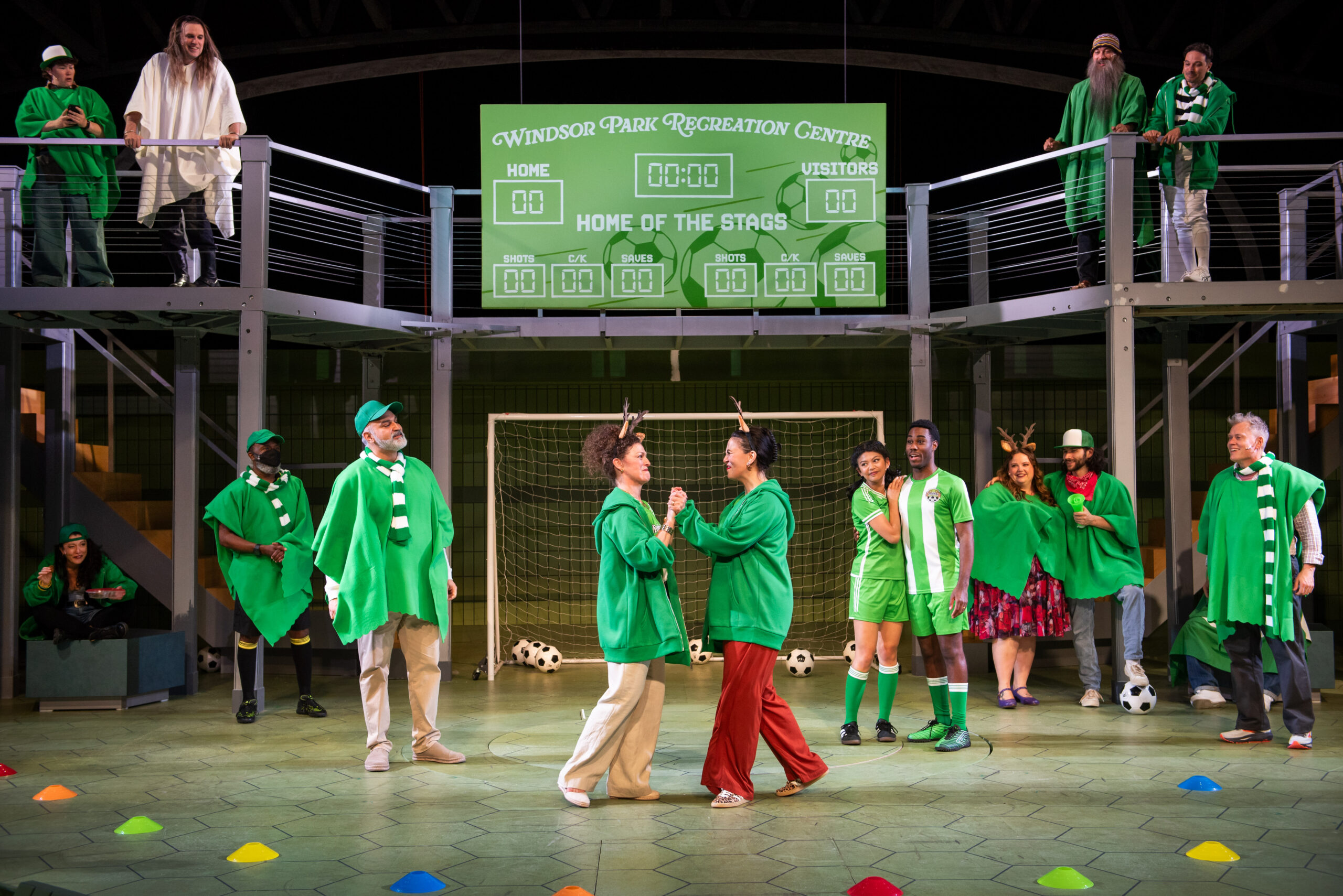 A diverse group of people in green sports attire stand in a recreation center, exuding excitement. The scoreboard reads "Windsor Park Recreation Centre."
