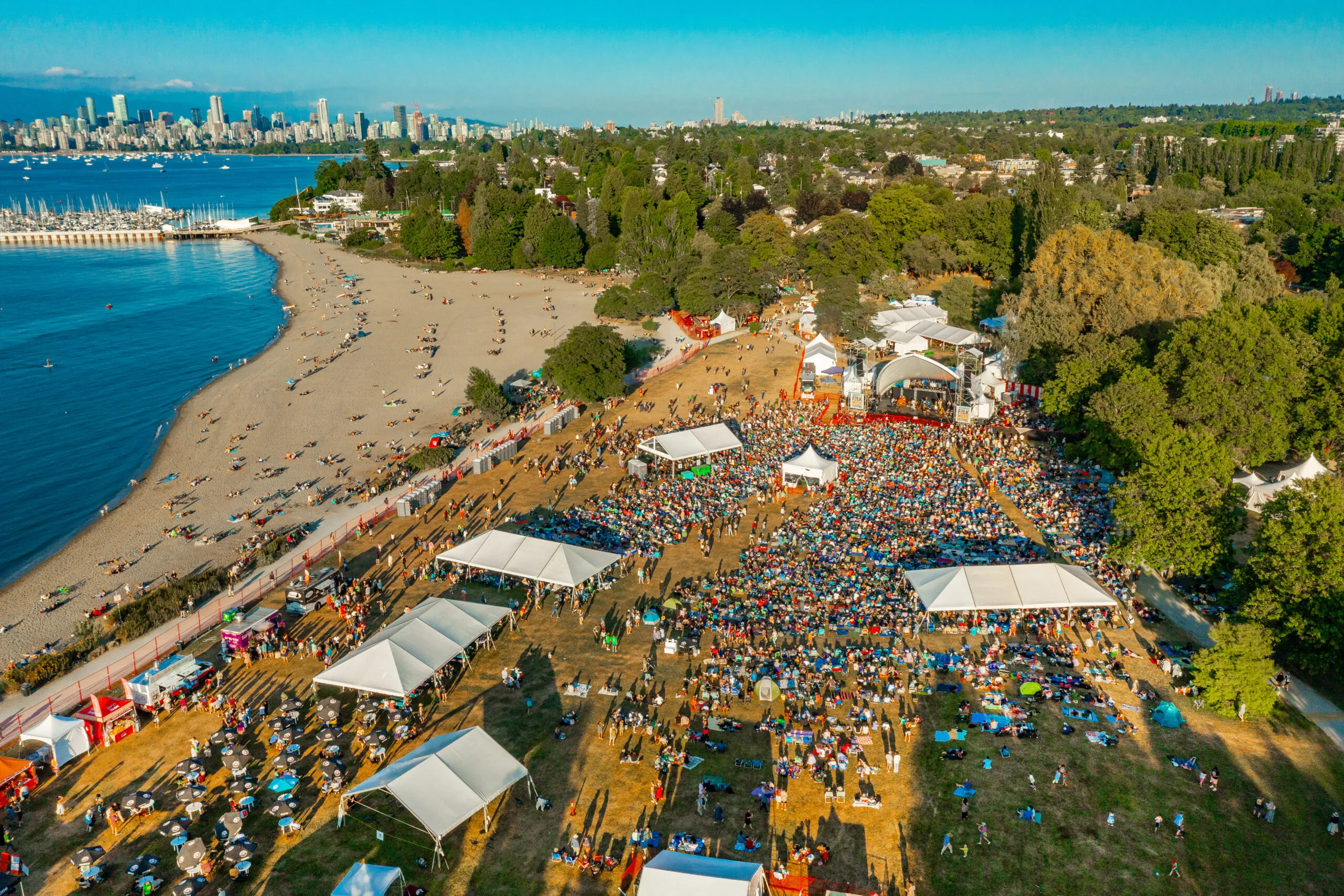 Aerial view of a large outdoor festival with crowds gathered around a stage. White tents dot the area beside a sandy beach, city skyline visible in distance.
