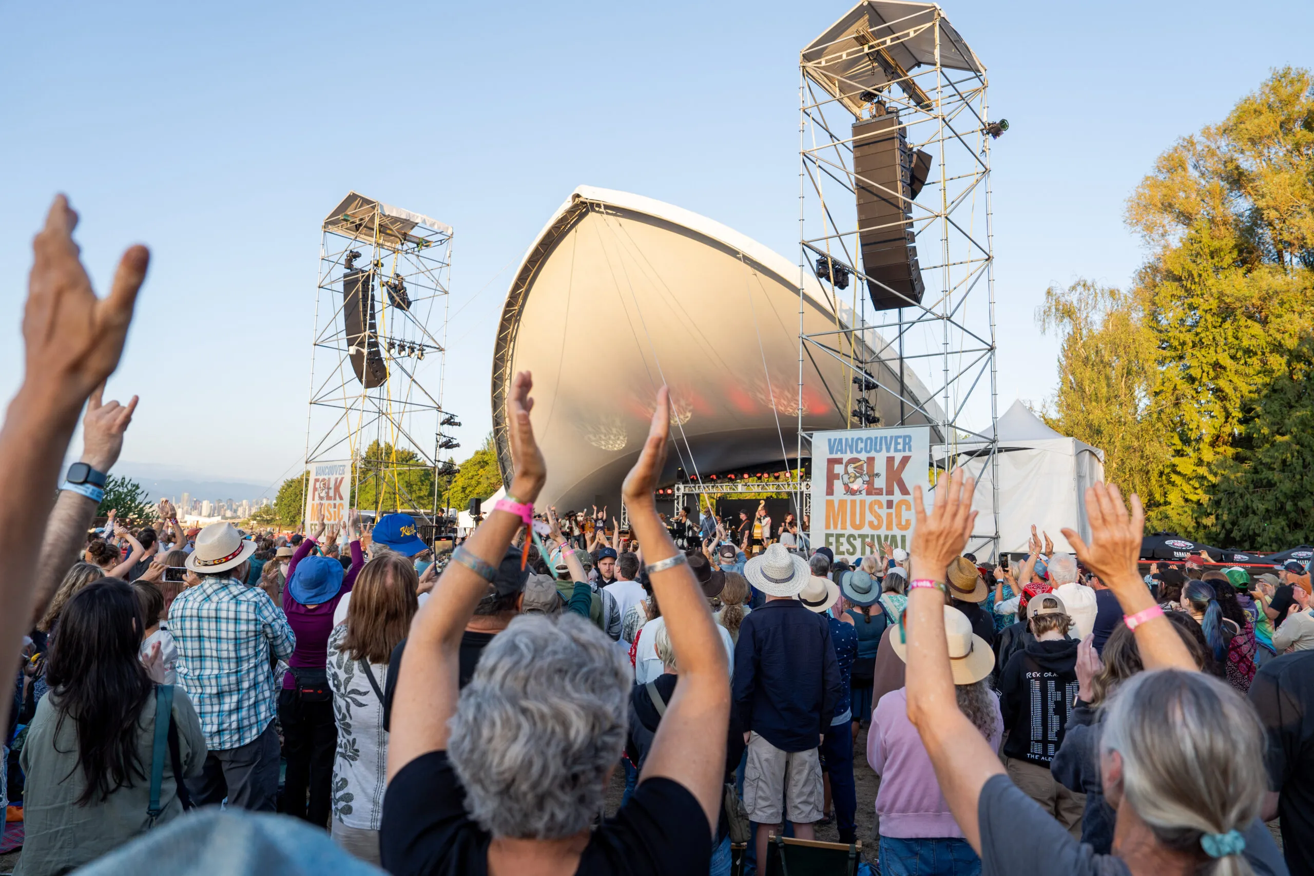 Crowd with raised hands cheers at an outdoor music festival near a large stage, marked "Vancouver Folk Music Festival," under a clear blue sky.