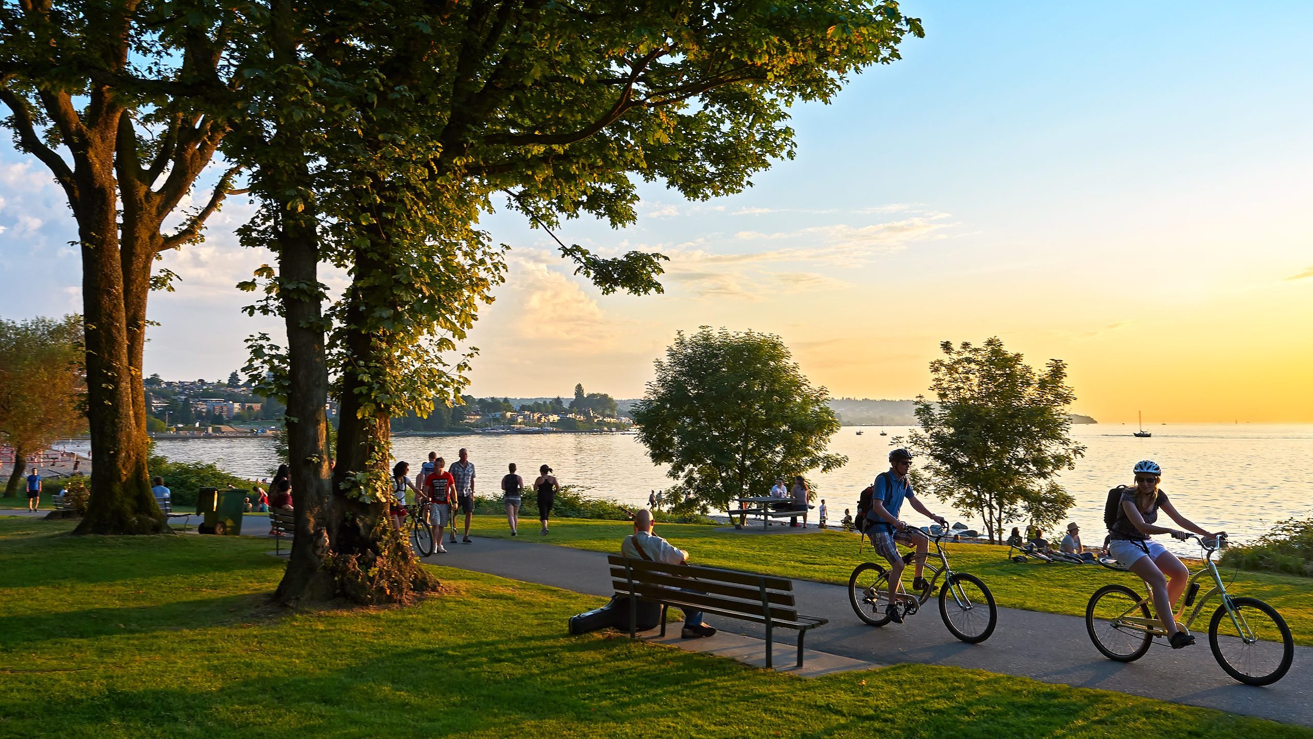 Cyclists ride along a park path at sunset, near a tranquil lake. People relax on benches and the grass, enjoying the serene, vibrant view.