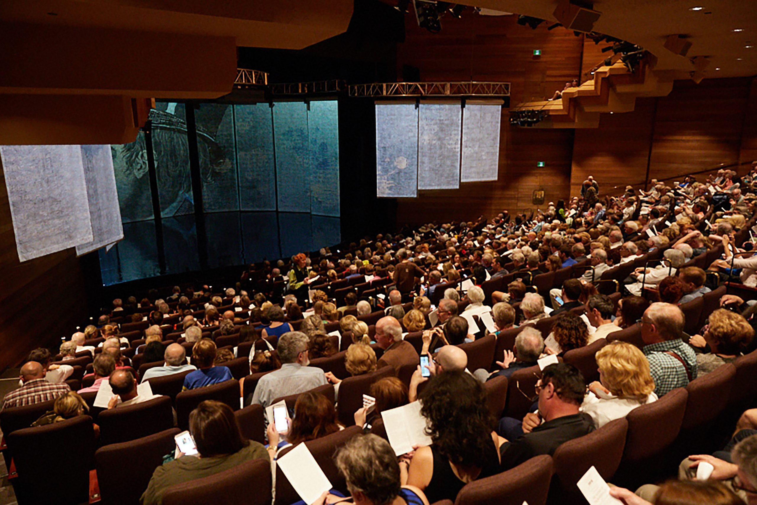 A large audience fills a theatre auditorium before a performance, with projected scenery displayed across the stage.