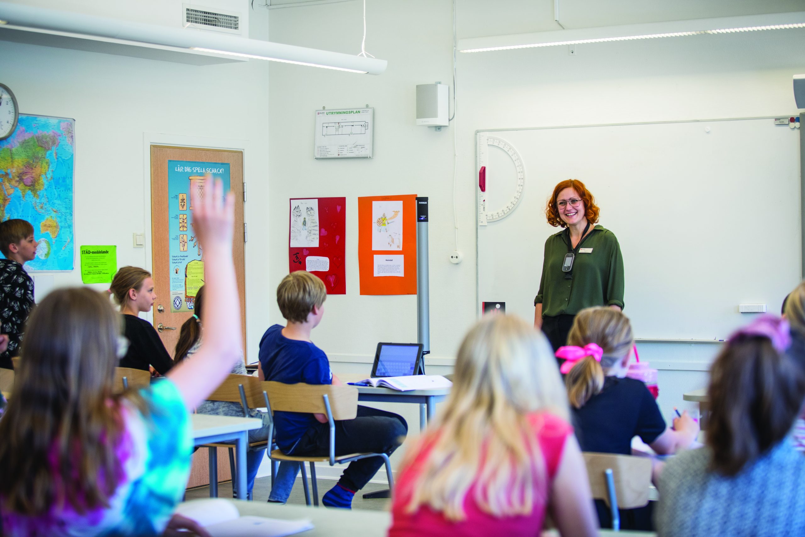A teacher wearing a wireless microphone smiles at the front of a classroom as a student raises their hand.