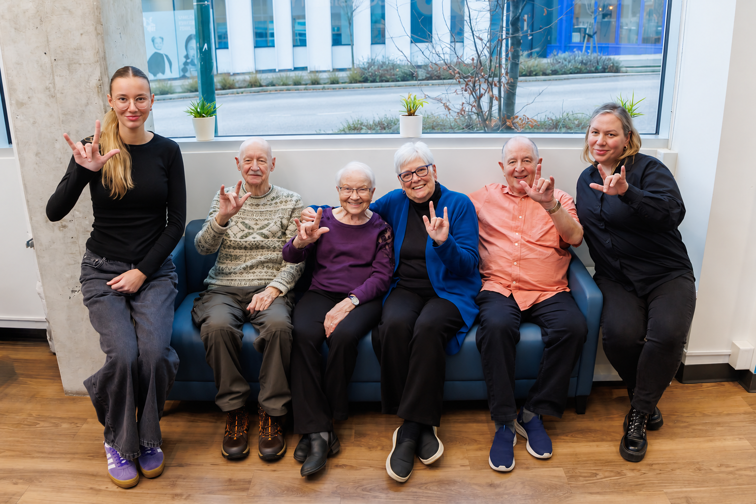 Six smiling people seated together on and beside a blue couch, each making the “I love you” sign in ASL.