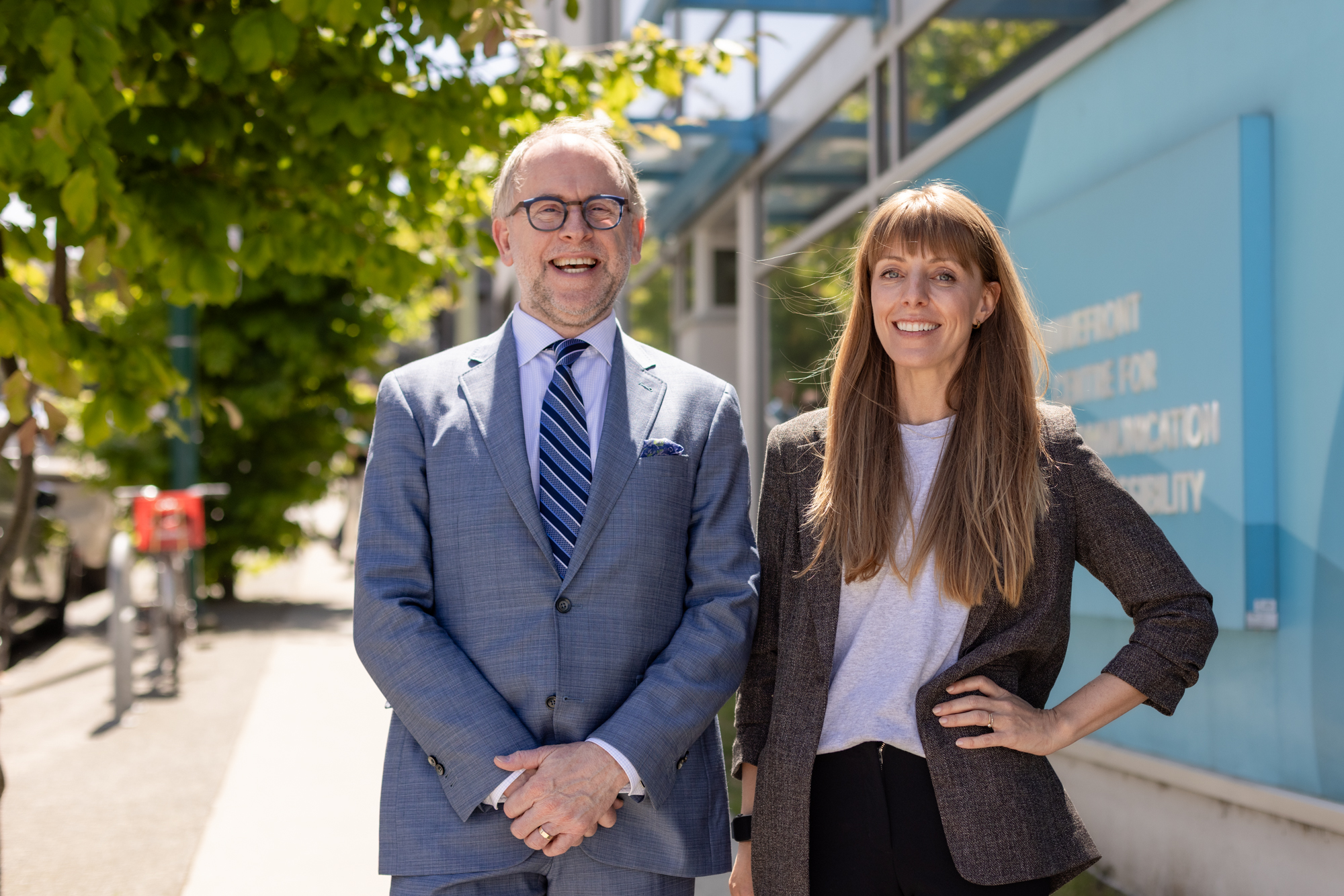 Two smiling people stand outside Wavefront Centre on a sunny day, with leafy trees and the building’s blue exterior behind them.