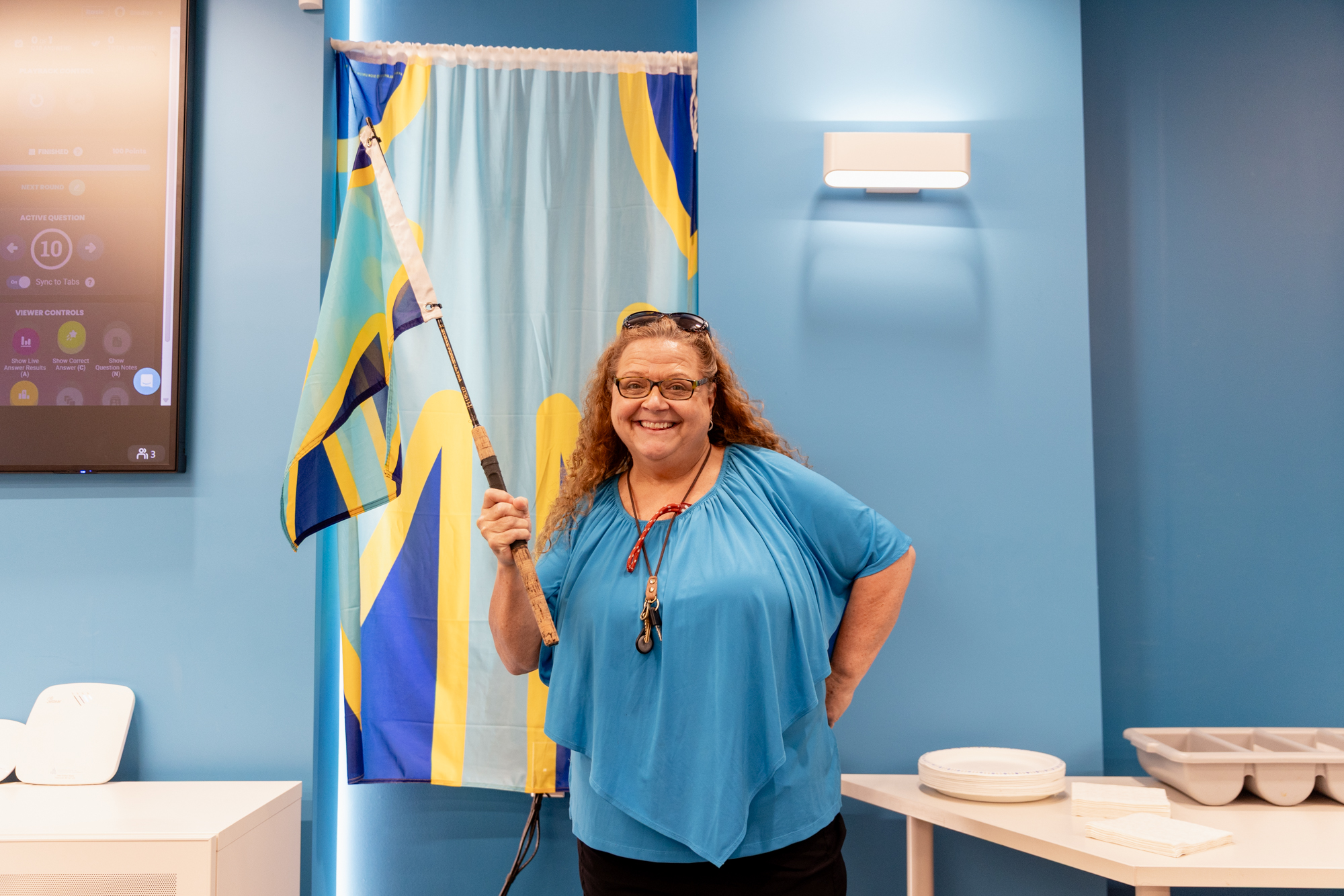 A smiling woman holds the blue, turquoise and yellow Deaf flag in front of a matching backdrop.