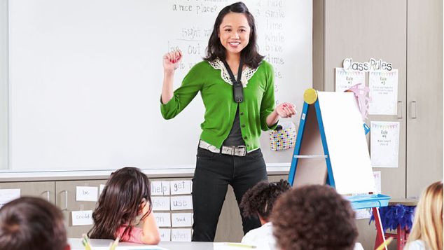 A smiling teacher wearing a wireless microphone leads a lesson as students work at tables in a classroom.