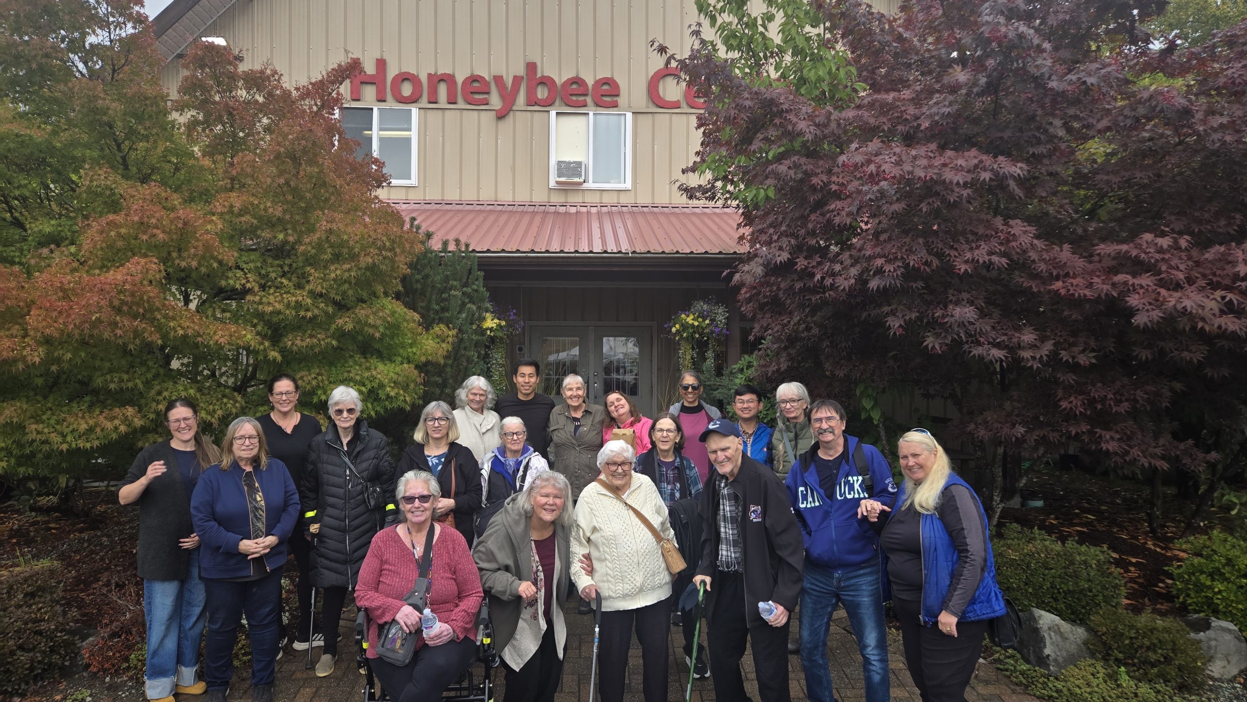 Participants in Wavefront Centre’s Better at Home Seniors Day outing pose outside the Honeybee Centre in Surrey, surrounded by trees with colourful autumn leaves.