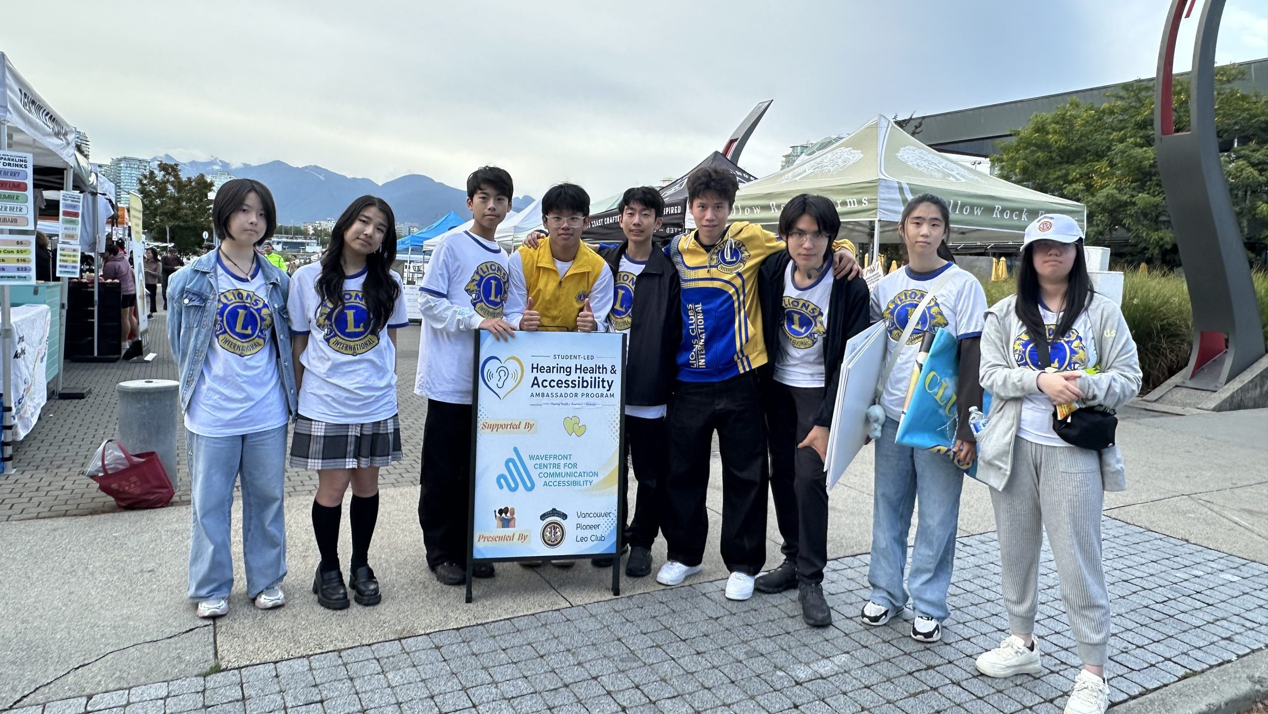 Nine Vancouver Pioneer LEO Club volunteers pose beside a Hearing Health and Accessibility Ambassador Program sign at the Vancouver Farmers Market in Milton Wong Plaza, Olympic Village. Market tents and mountains are visible behind them.
