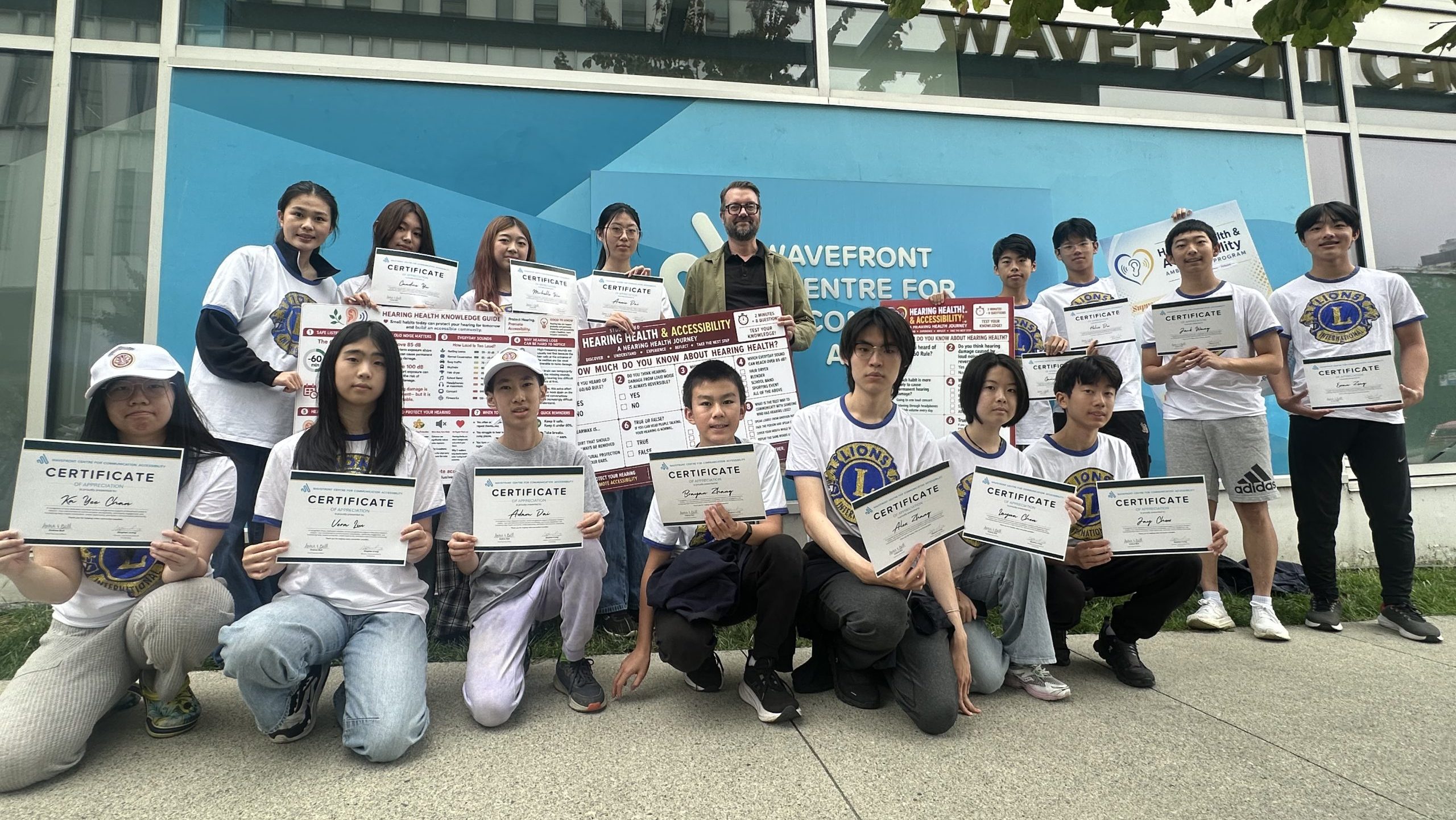 Stephen Irving, Director, Communications & Fundraising, poses outside Wavefront Centre with Vancouver Pioneer LEO Club students holding volunteer appreciation certificates and hearing-health posters in September 2026.