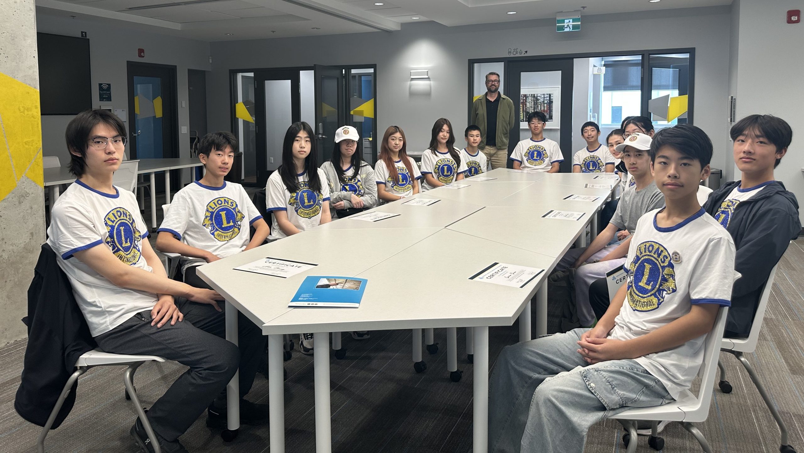 Vancouver Pioneer LEO Club students wearing Lions International shirts sit around a conference table at Wavefront Centre, with certificates and program materials in front of them. An adult stands behind the group.