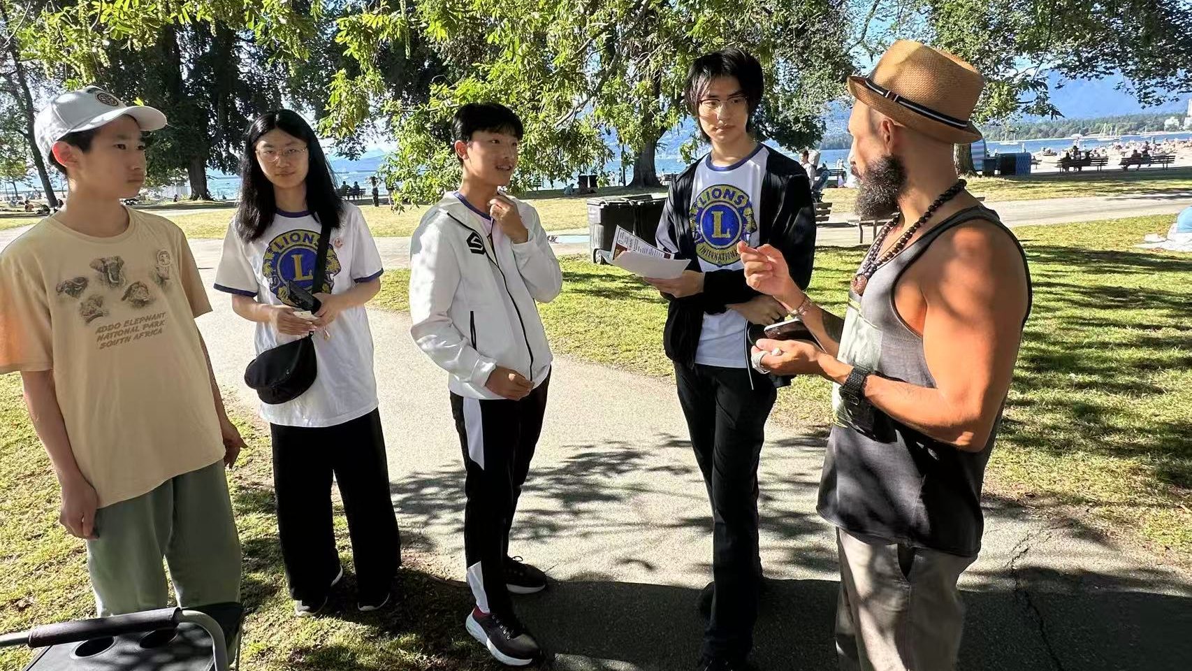Four Vancouver Pioneer LEO Club student volunteers speak with a community member during hearing-health outreach at Kitsilano Beach. One volunteer holds educational materials, with trees, the beach and ocean behind the group.