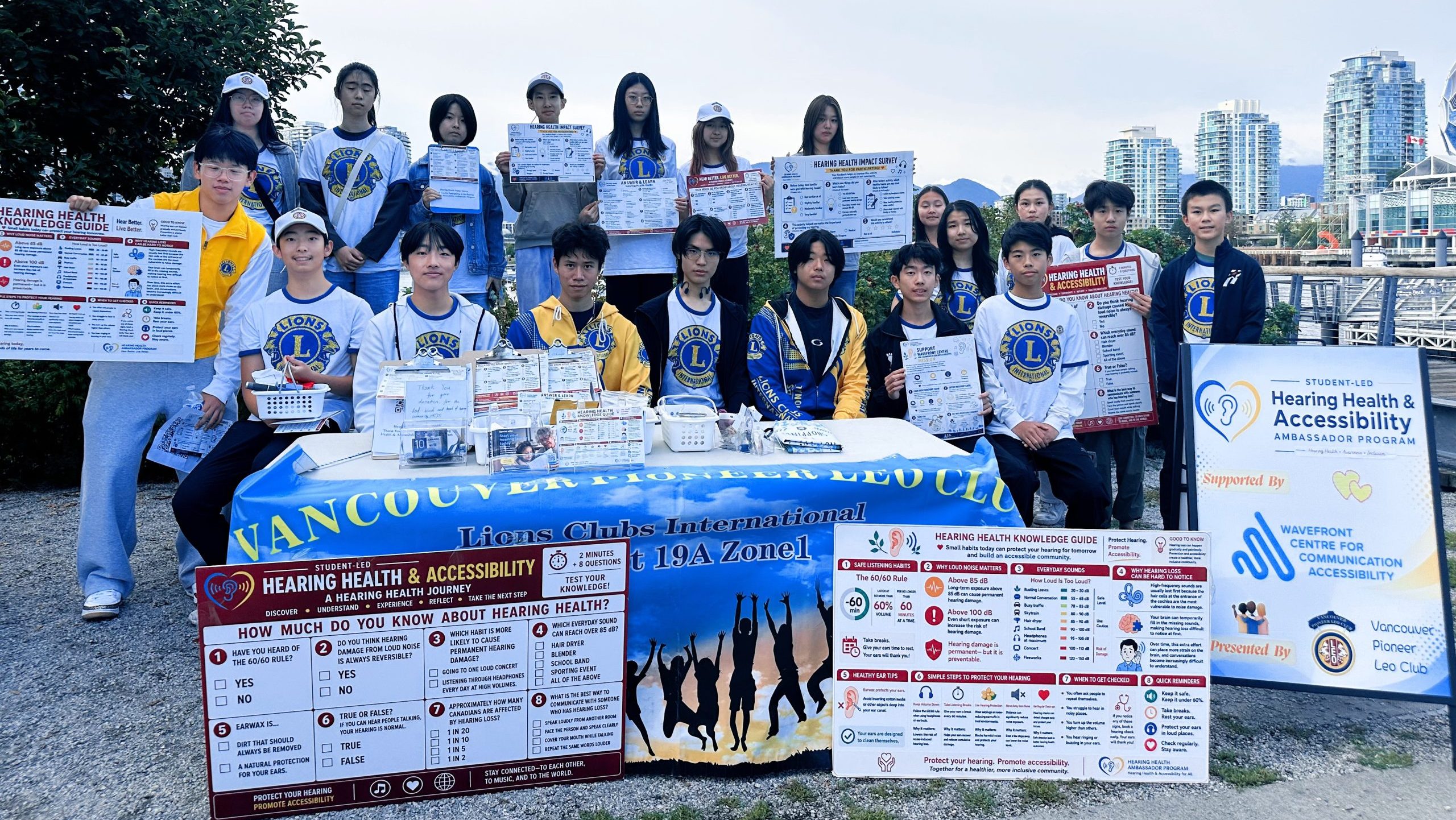 Vancouver Pioneer LEO Club student volunteers pose behind a hearing-health outreach table in Olympic Village, holding educational posters and surveys. A sign recognizes Wavefront Centre’s support, with False Creek and waterfront buildings in the background.